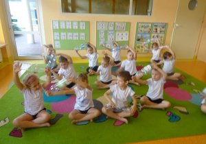 Dzieci siedzą skrzyżnie z uniesionymi nad głową rękami w których trzymają plastikowe butelki, wykonują skłony na boki.